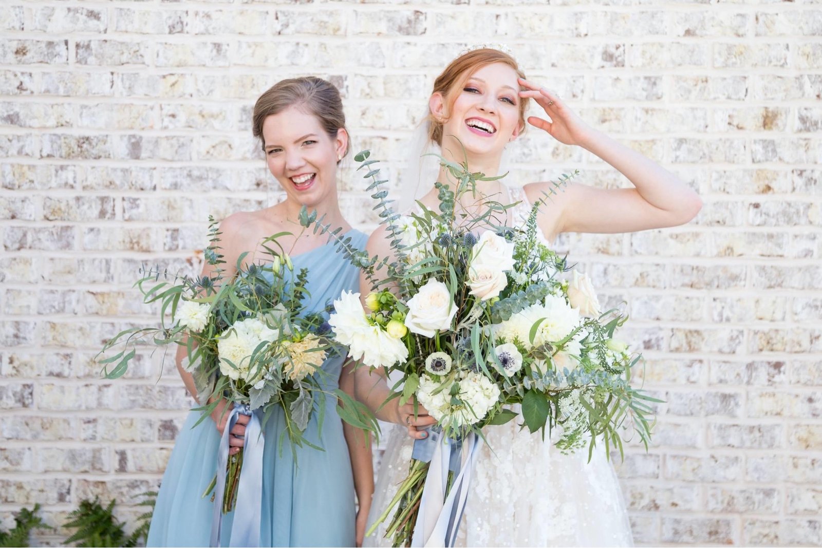 Bride and bridesmaid laughing with bouquets against a brick wall
